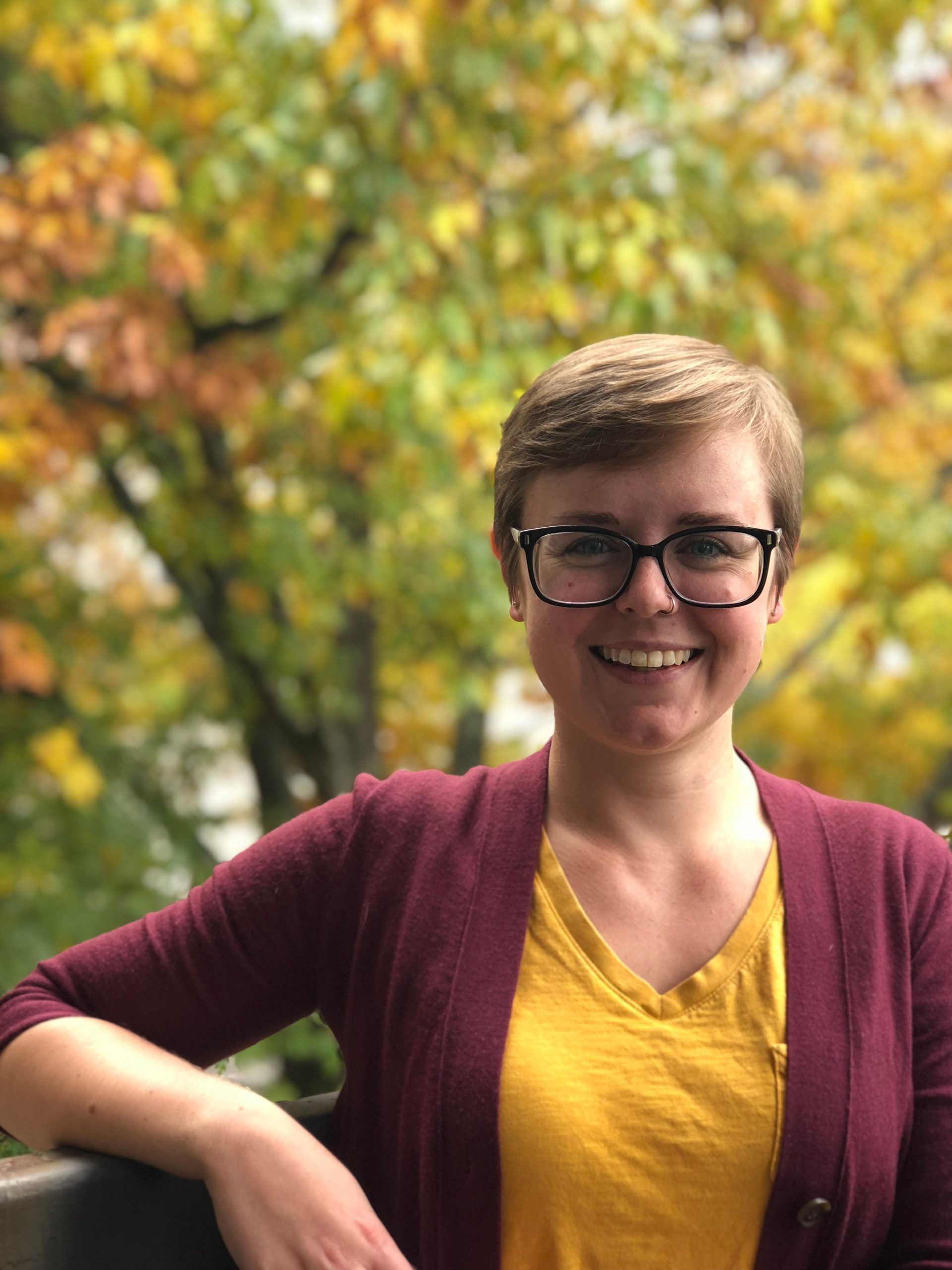 woman with short hair and glasses with her right elbow on a railing. a tree with green, yellow and red foliage fills the background