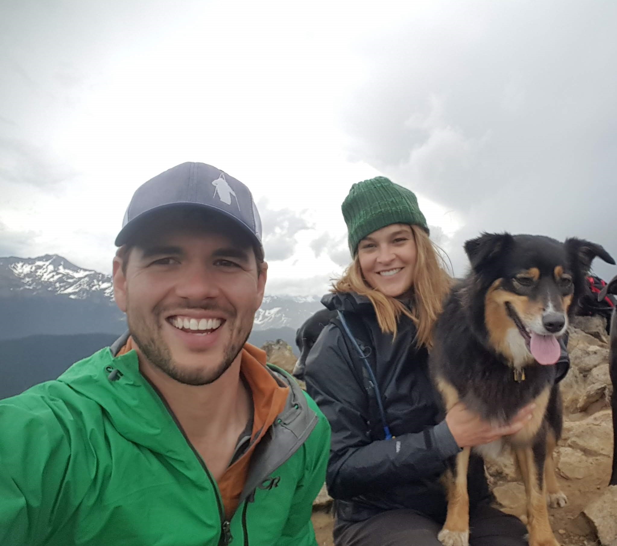 a man, a woman and a really cute dog with snow-covered mountains in the background