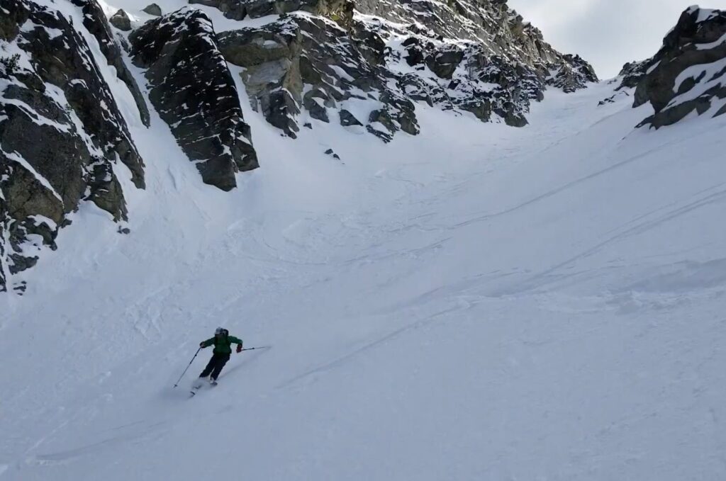 man skiing down the face of a backcountry mountain with craggy black rocks in the background.
