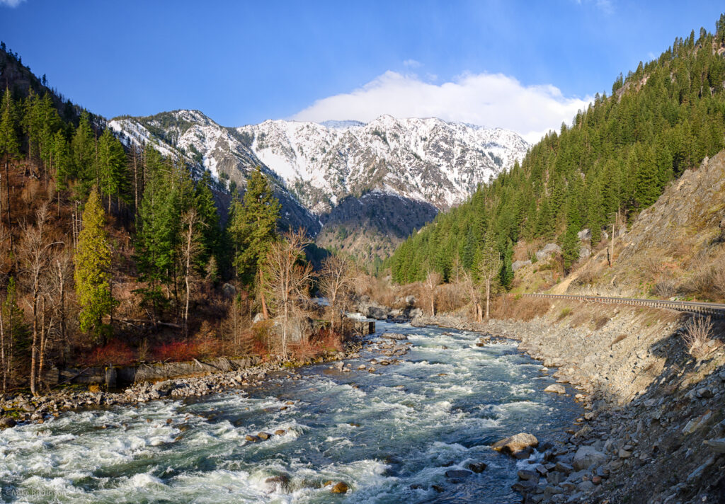 river between two wooded hills with snowy mountains in the background.