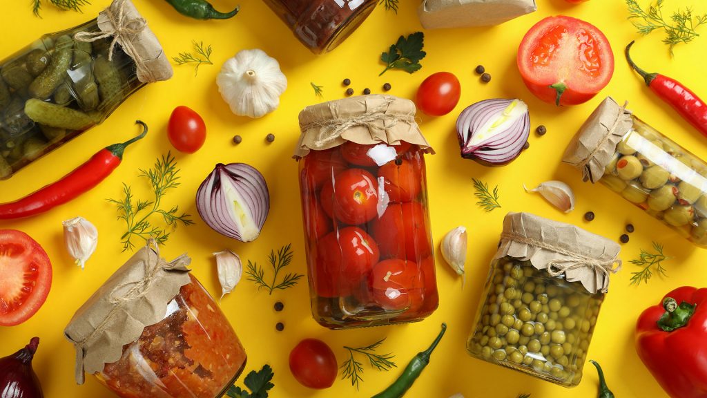 A bright yellow background with various different types of canned food in mason jars, including tomatoes, capers, and peppers. Around the canned goods are a wide variety of vegetables and spices strown about, including, tomatoes, peppers, garlic, onions, peppercorns, dill, and parsley.