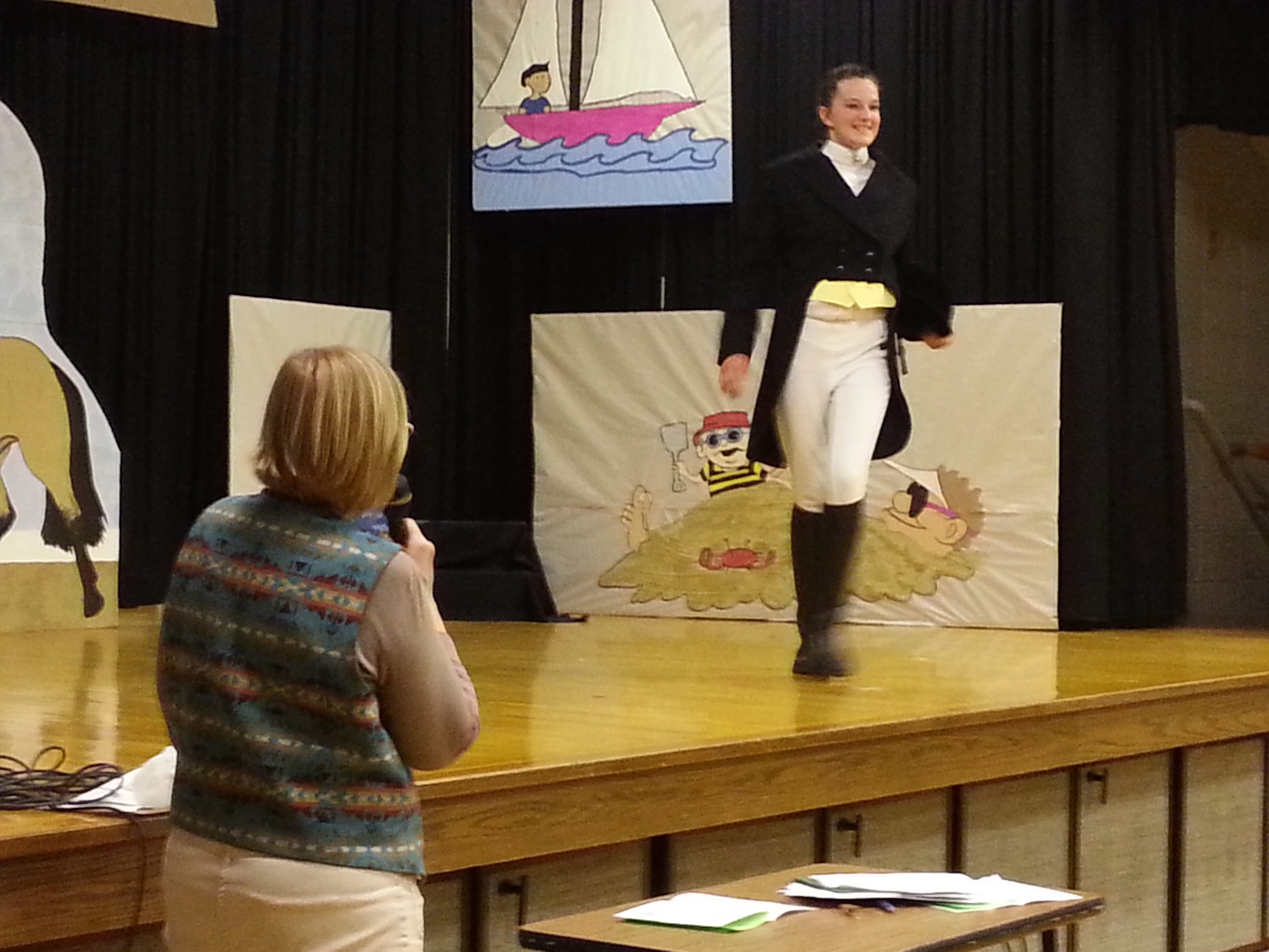 Youth in a black tails coat, white trousers, and boots models on stage while an adult narrates