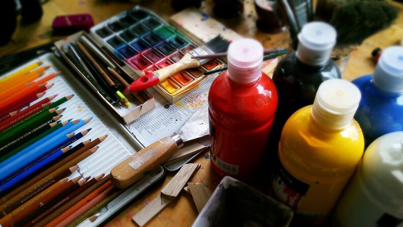 A selection of colored pencils, paint brushes, watercolors, bottles of paint, and other art supplies on a wood table.