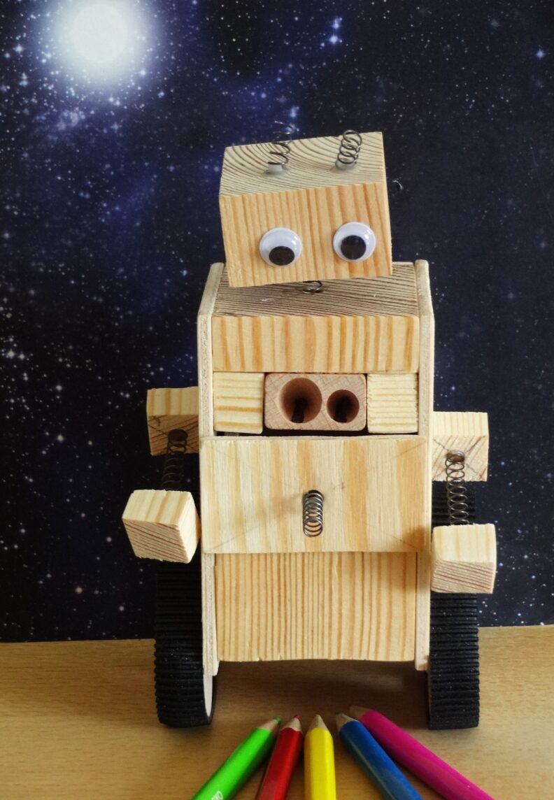 A wooden robot with googly eyes with a simple pencil sharpener built into its chest looks down at a pile of colored pencils.