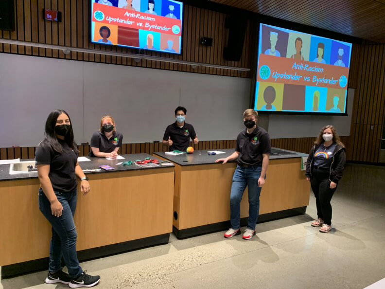 Five youth present at a conference, their presentation title on the slide show behind them reads "Anti-Racism: Upstander vs. Bystander". There are two tables iwht their presentation materials at the front of the room, two youth stand behind it, three in front