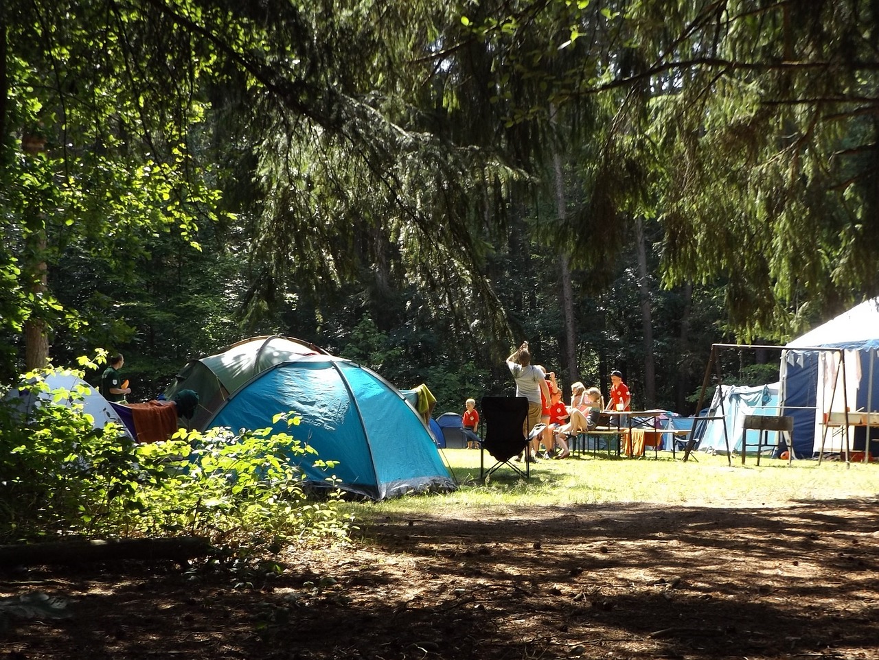 A campsite in the woods with multiple tents, picnic tables, and chairs. There are a number of people in the distance at the campsite