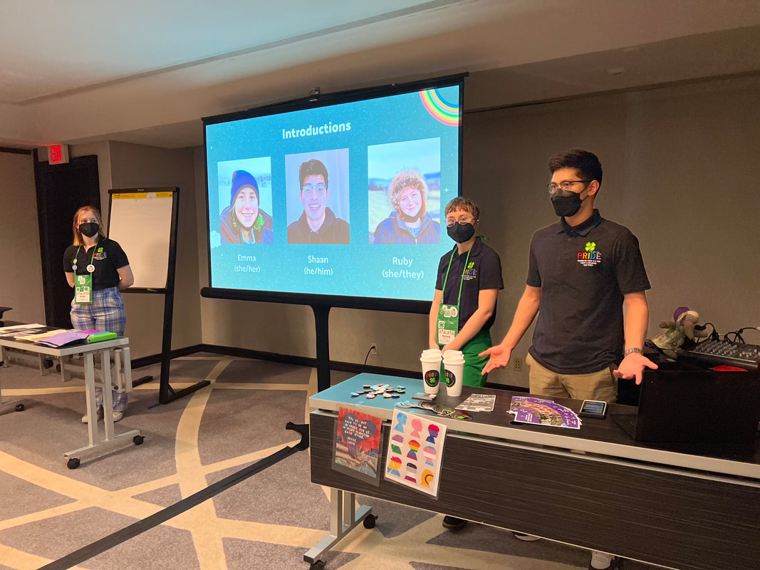 Three youth stand while presenting in a conference room. They have tables in front of them with their documents and fliers and on the screen behind them are their photographs on an introduction slide