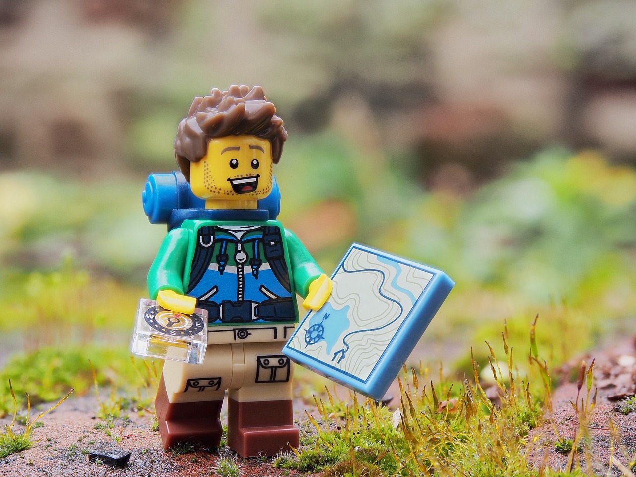A LEGO minifigure in hiking atire holds a map tile and compass tile. The minifigure stands on a rock with moss on it