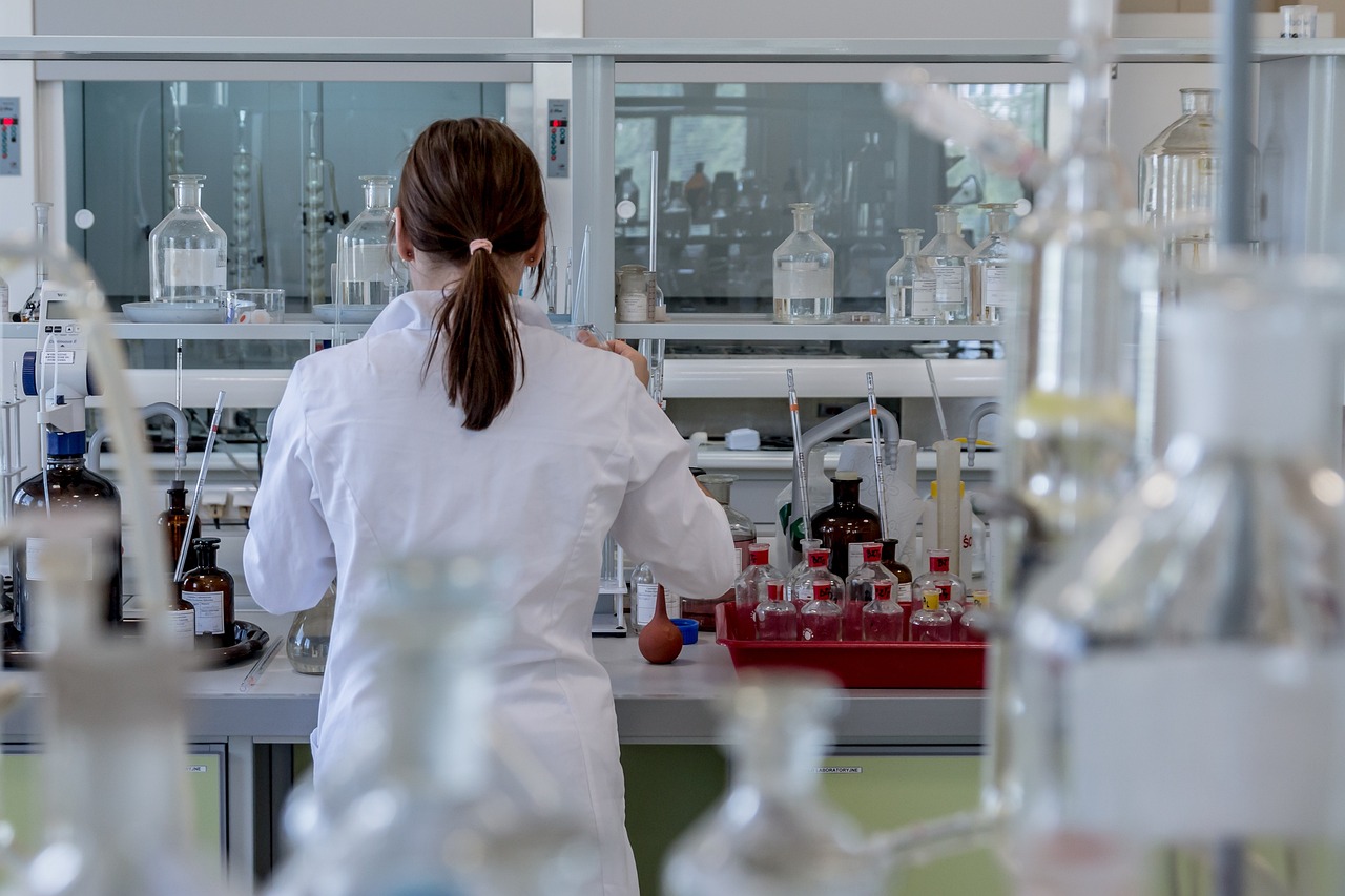 A person with their hair in a ponytail stands at a lab counter with their back to the viewer. They are surrounded by chemists' equipment