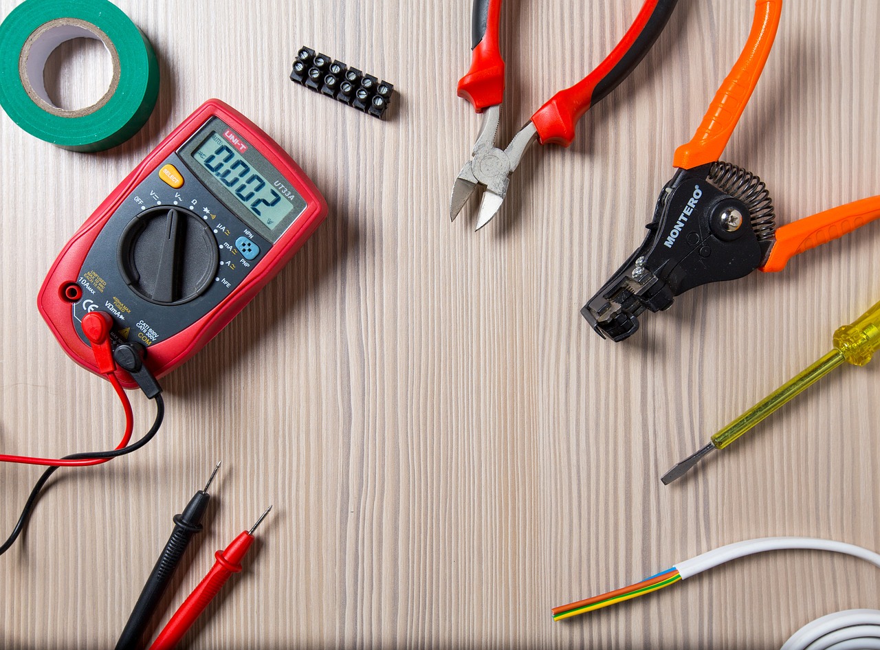 A multimeter, electrical tape, wire cutters, wire strippers, a screwdriver, electrical wiring, and connectors on a wood table