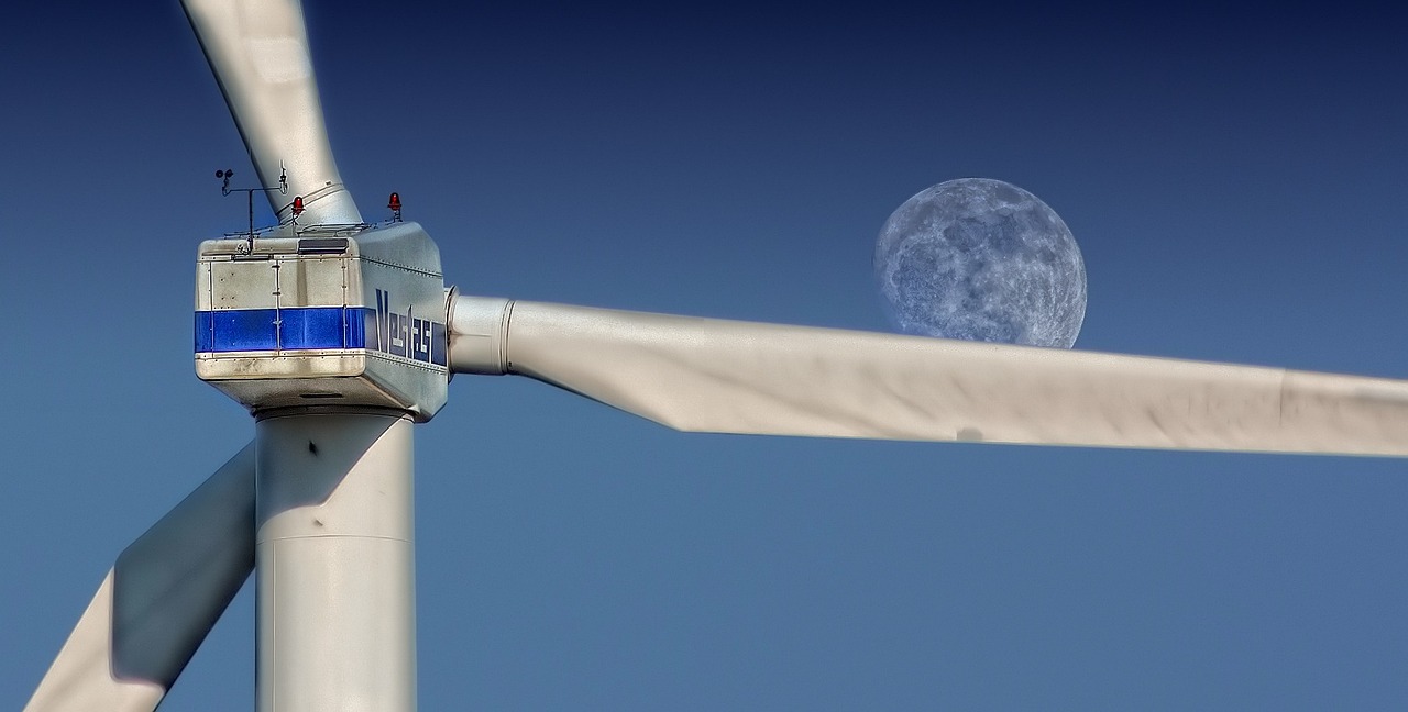 Close up of the head and blades of a large wind turbine with the moon visible over one of the turbine blades
