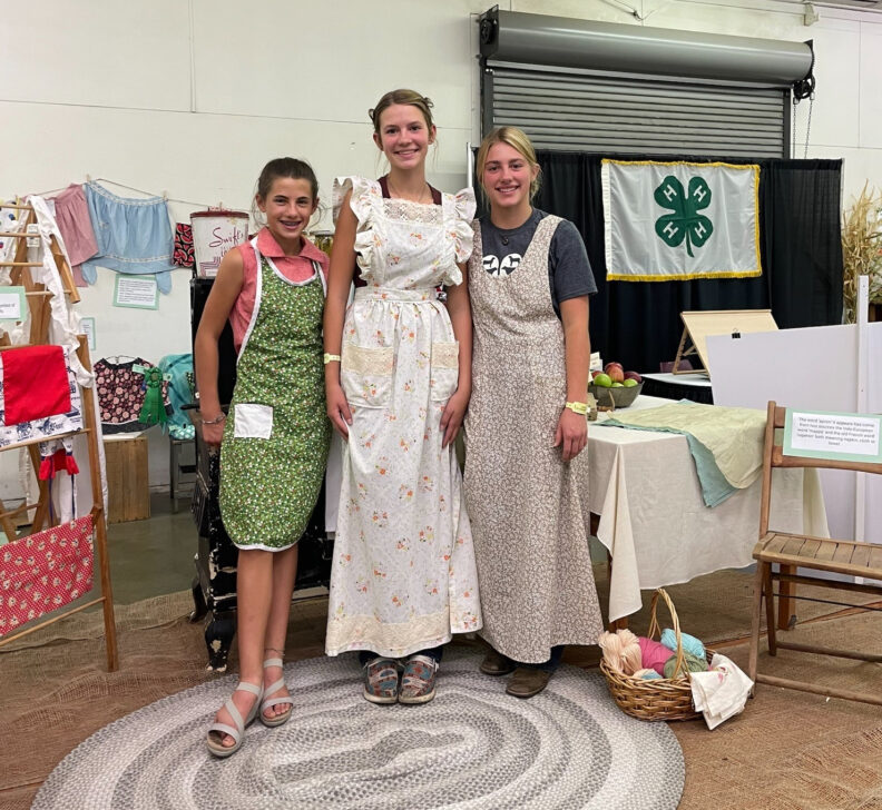 Three youth wearing long full front coverage aprons , one green and knee length, two floor length in white and tan
