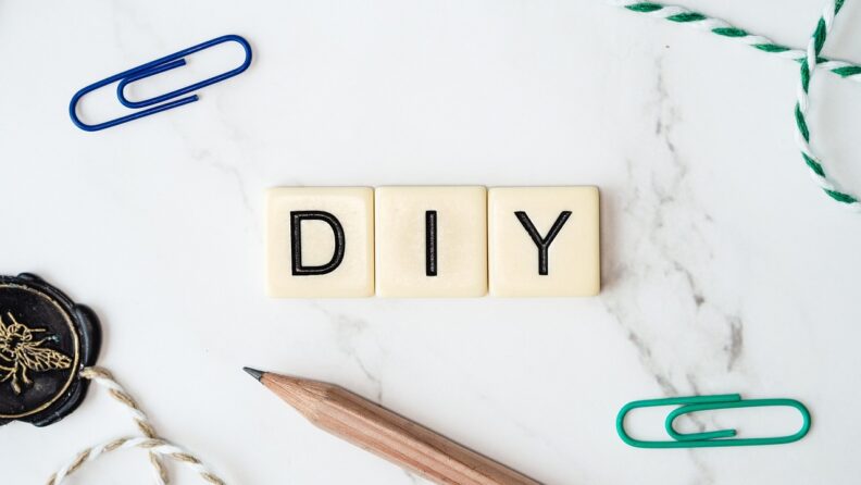 Paperclips, a pencil, string, and a black container with a bee on it sit on a marble tabletop with the letters DIY on plastic tiles at center