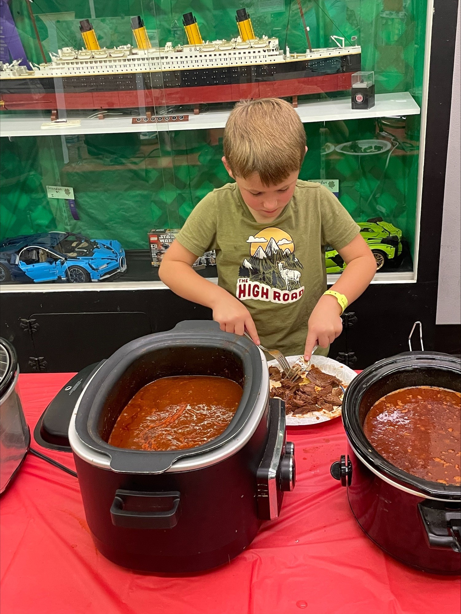A youth prepares meat during a chili cooking contest while standing behind crock pots on a red table. There are model cars and a model of the Titanic on a shelf in the background
