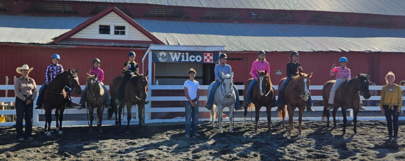 Seven youth on horses outdoors in front of a barn, three individuals stand to the sides and in the middle.