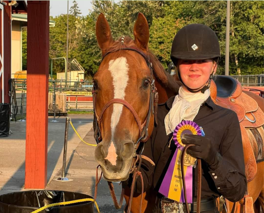 A youth stands beside her horse in her riding outfit holding a yellow and purple award ribbon.
