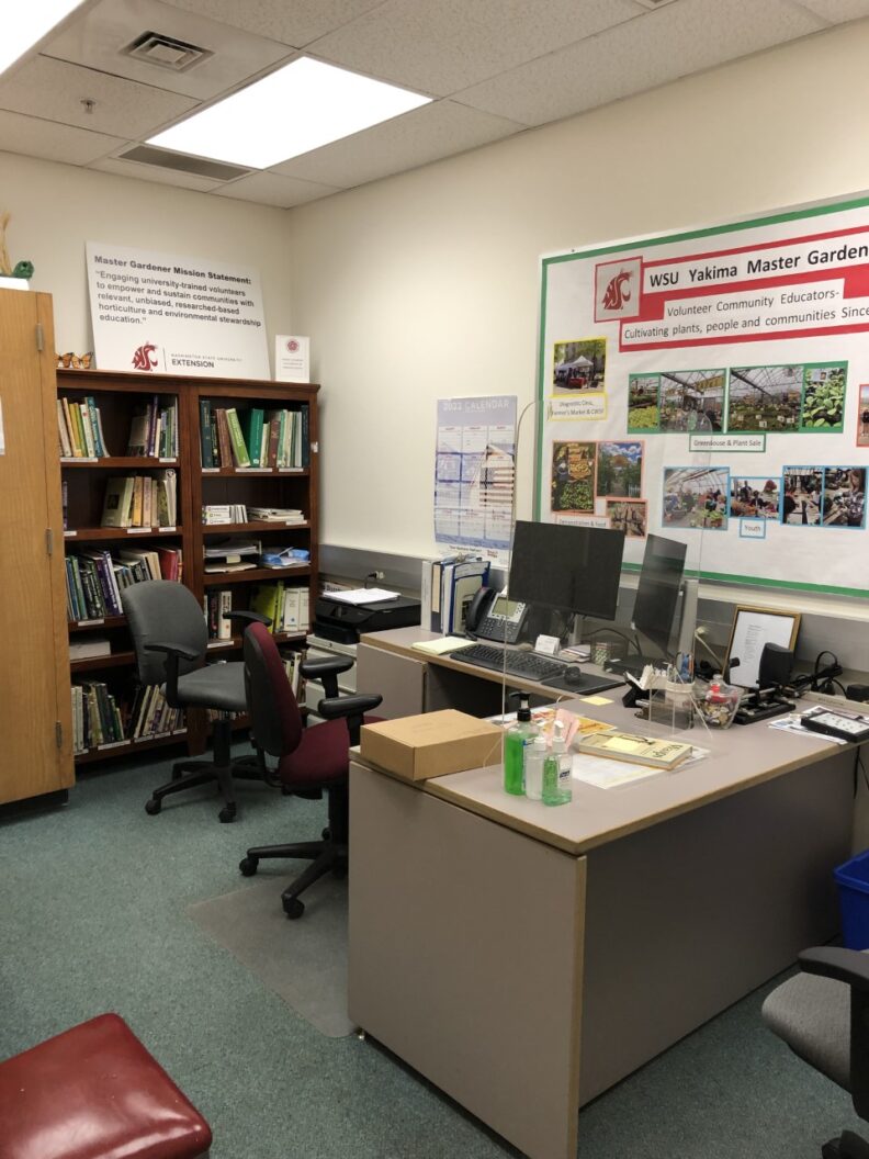 The plant clinic office with rolling chairs around an L shaped desk.  There is a computer on the desk with two screens and a bulletin board for the Master Gardeners behind the desk.  A bookshelf of resources is at the back of the room.