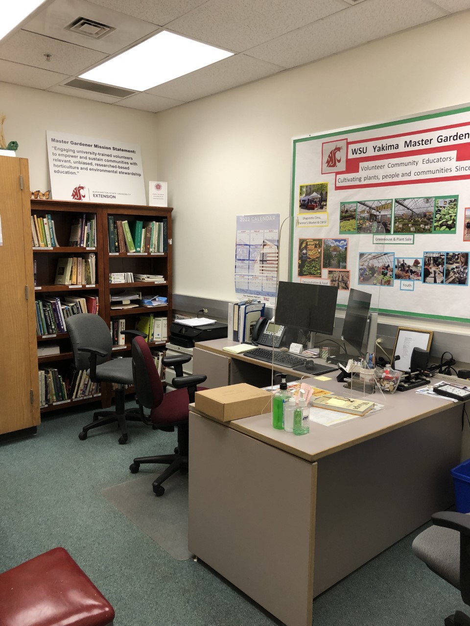 The plant clinic office with rolling chairs around an L shaped desk. There is a computer on the desk with two screens and a bulletin board for the Master Gardeners behind the desk. A bookshelf of resources is at the back of the room.