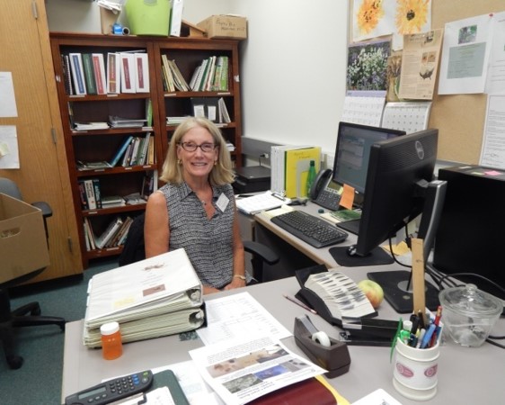 A smiling master gardener volunteer sits at an L shaped desk.  She is facing the camera, reference material on the desk surface between her and the viewer.  To the viewer's right, there is a computer with two monitors.  Behind the master gardener, there is a shelf of reference material.