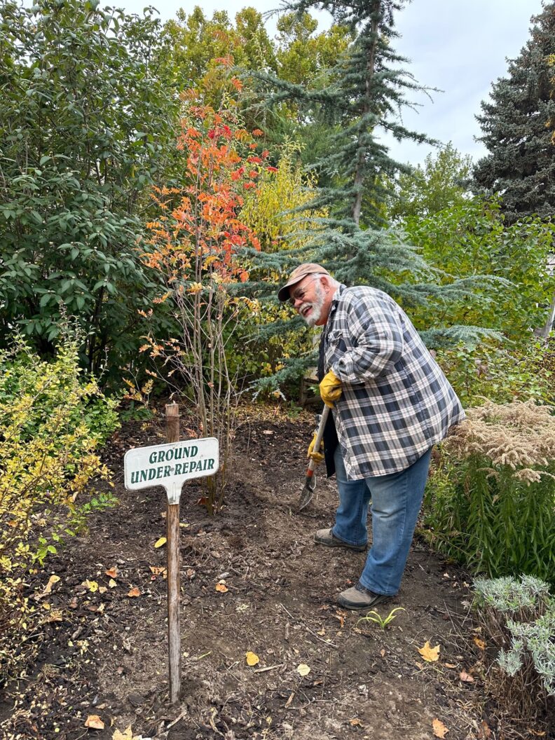 A volunteer wearing a hat, jeans, and flannel shirt works the soil beside shrubs and trees near a sign that reads "ground under repair."