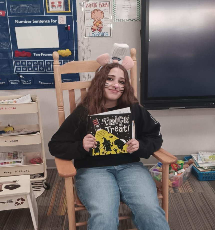 A teenager with whiskers painted on her cheeks and a tiny chef's hat on her headband holds up a Halloween book while sitting in a rocking chair in an elementary school classroom.