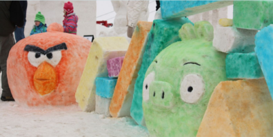 A close-up of Angry Birds characters and blocks built out of colorfully dyed snow. At left, a large reds cranky round bird and at right, a large round green pig-like orb. There are people milling in the background, enjoying the sculpture.