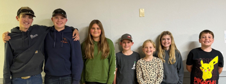 Seven youth standing against a beige wall.  They are smiling, the two far left young people have their arms around one another.