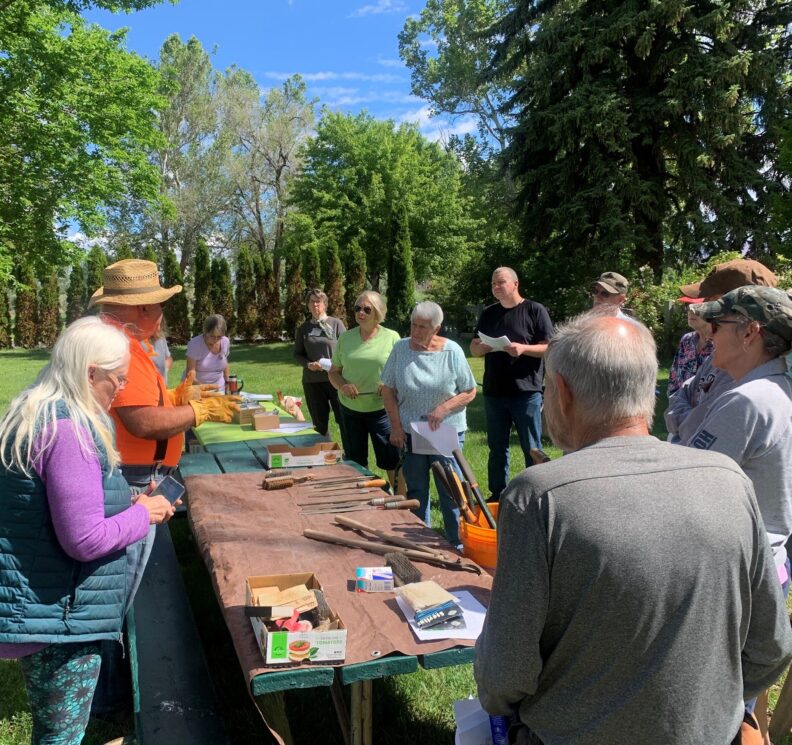 Master Gardeners present to a group of participants outdoors.  Their tools and presentation materials are spread out on a tarp over a green picnic table.  The grassy area of the park is surrounded by a variety of trees.