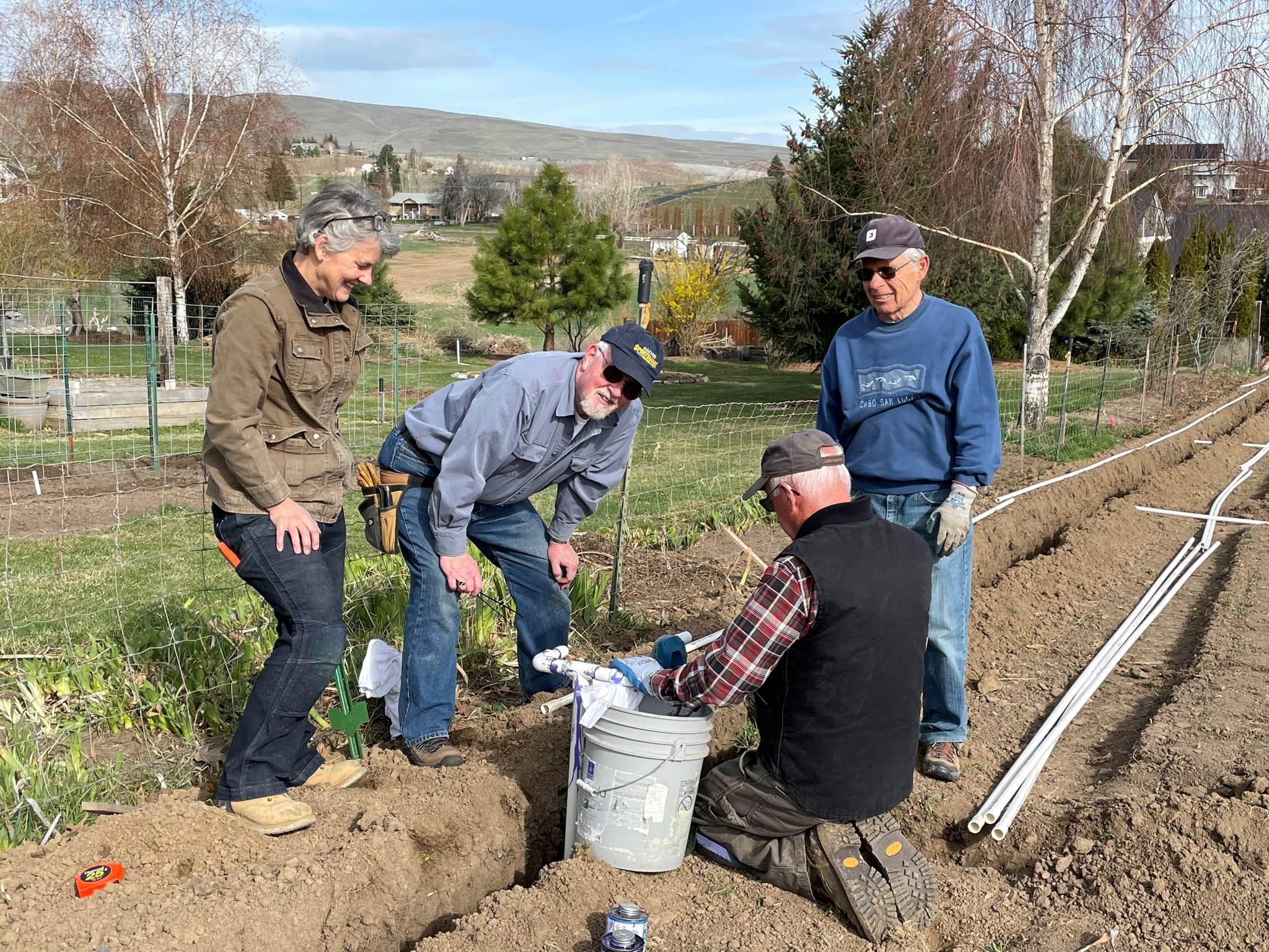 Four garden volunteers around a trench dug in the dirt.  There are white PVC piples laid out on the dirt beside the trench.  One of the volunteers is preparing the pipes while three others assist. 