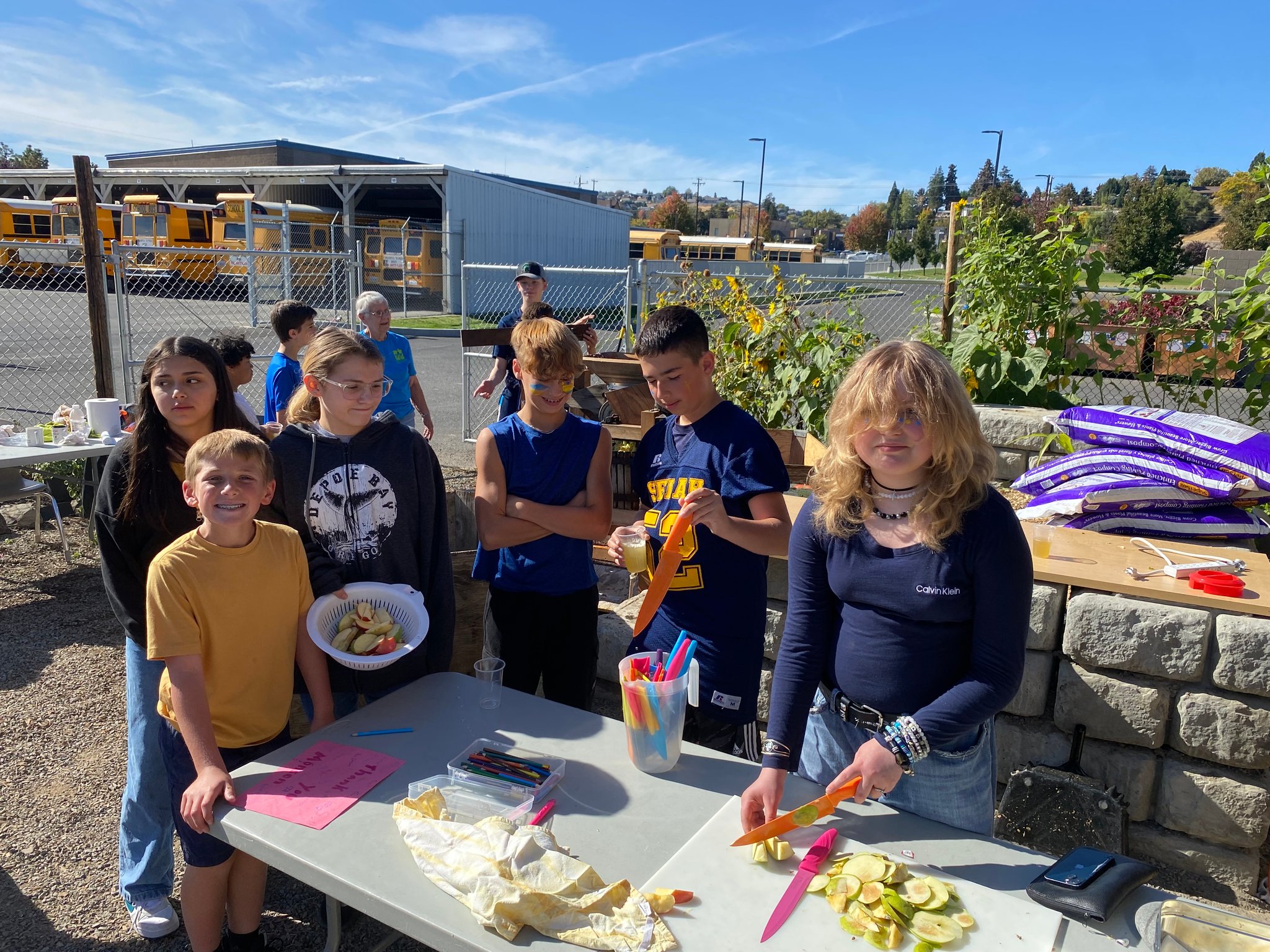 A group of six youth stand around a table.  One young person on the left is holding a colander of apple slices, one young person selects a knife for the task, and another youth is cutting apple slices.  They are outdoors in a garden surrounded by a tall chain-link fence.  There is a bus garage in the background.