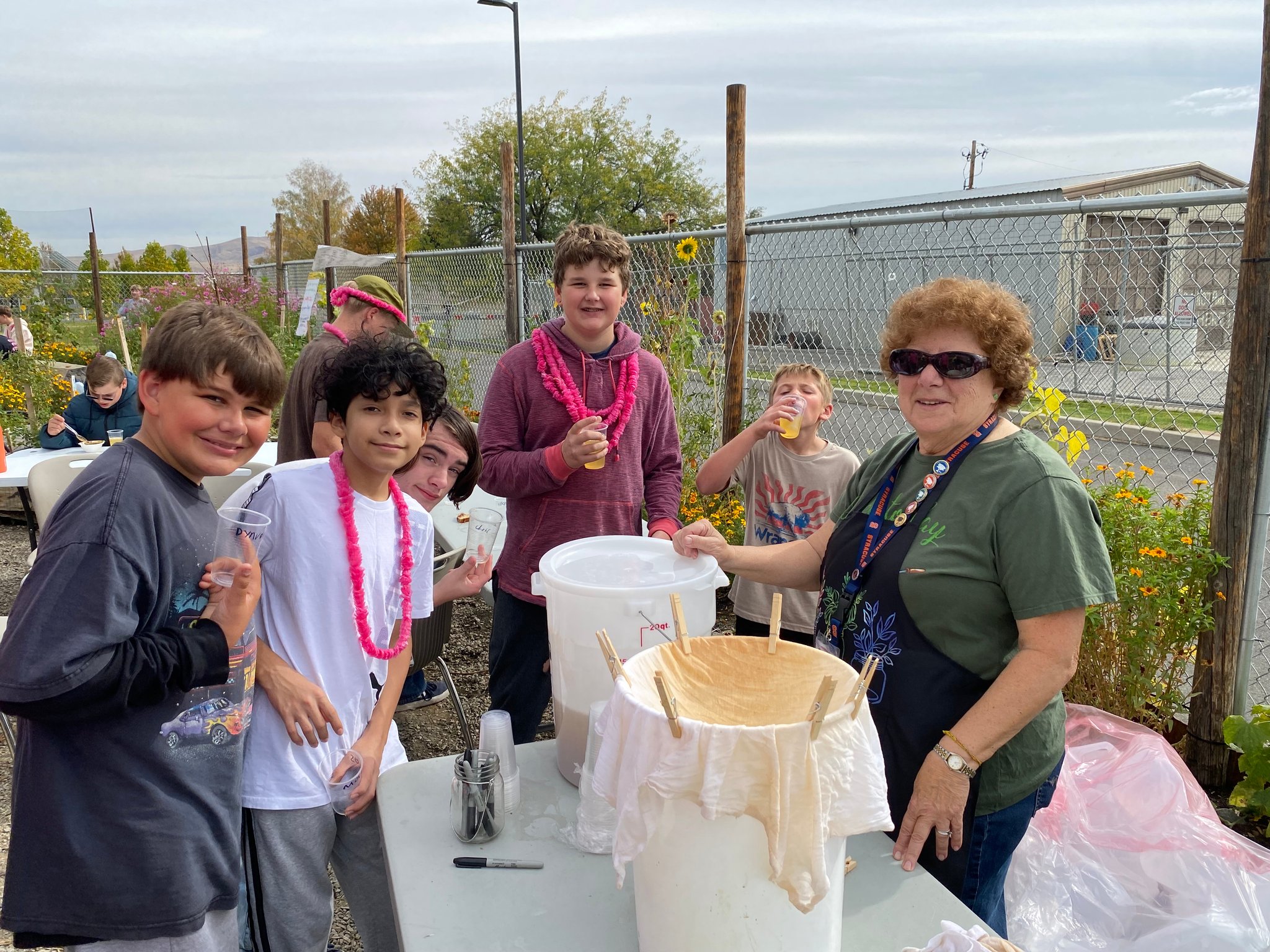 A group of older children and young teens gather around a table with a Master Gardener volunteer sampling fresh cider that has been strained in a bucket on the table.