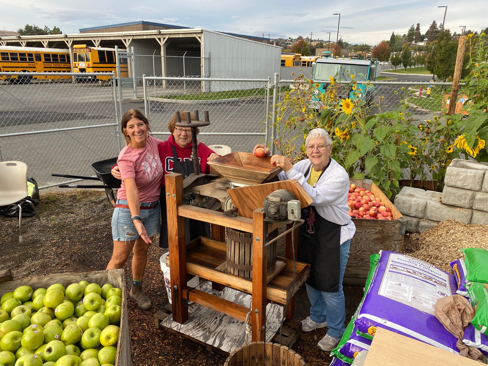 Three Master Gardeners show off the cider press.  At left, one has their arm around the other person, who is gesturing to the press.  On the right, another volunteer looks at the camera while holding an apple above the hopper that feeds apples into the press.