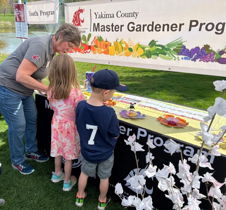 A Master Gardener bends down to work with two children visiting the program's outdoor table.  There are samples of a flower craft on the table and informational materials.