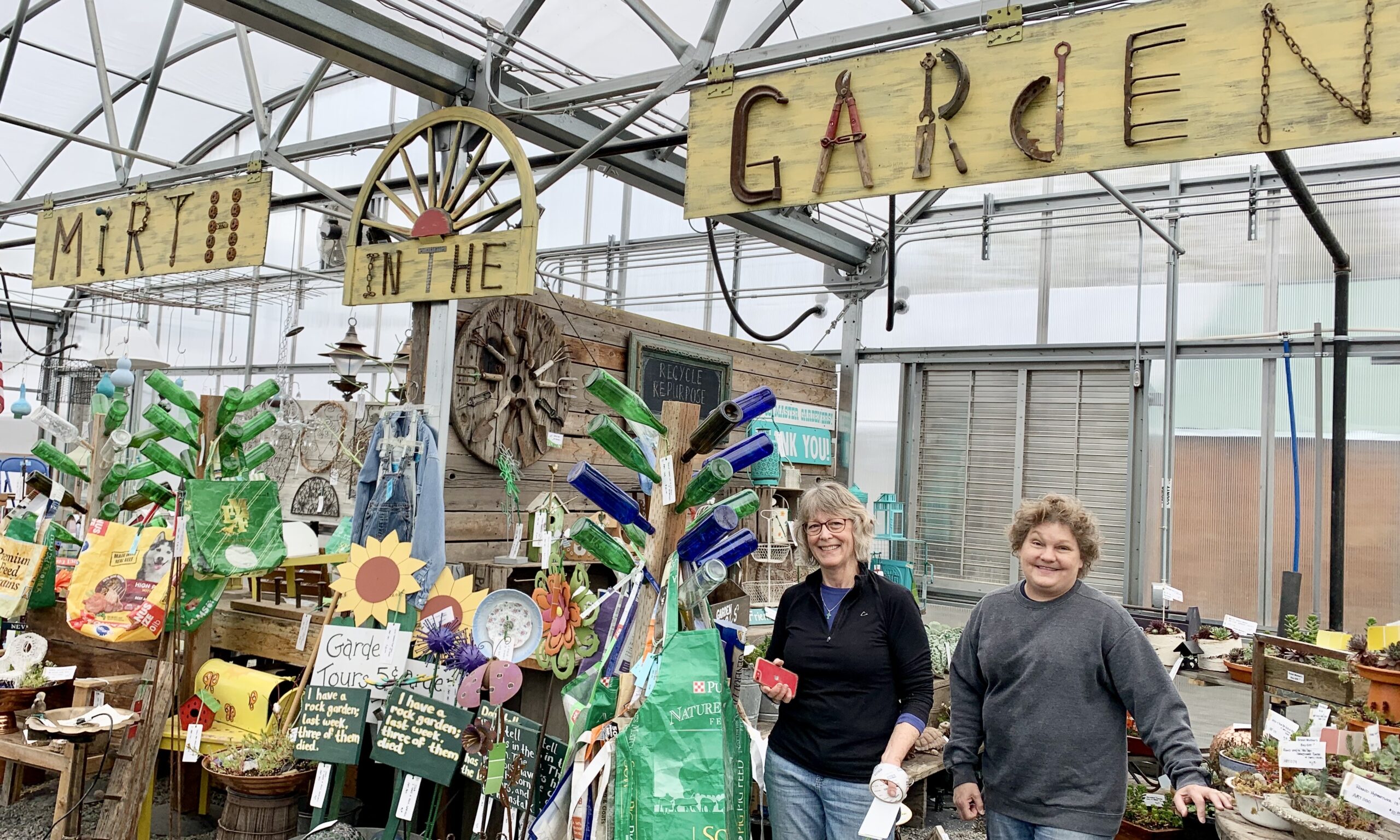 Two Master Gardener volunteers stand beside a collection of garden decoration for sale, such as bottle trees, wooden signs with flowers on them, and metal decorations. They are under a sign made from wood boards and old tools that spells out the phrase, "mirth in the garden."