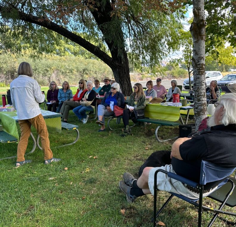A Master Gardener presents to a large audience of participants sitting two trees on a grassy lawn. The Master Gardener has materials spread out on a picnic table with a green plastic table cloth. Some participants sit in folding camp chairs while others are at the picnic tables. There is a parking lot in the background.
