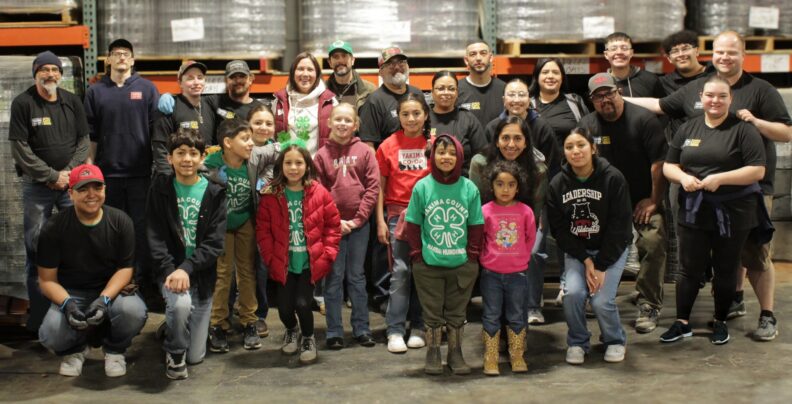 Large group of adults in black shirts smiling and standing behind a group of smiling youth members in green Yakima County Harrah Humdingers 4-H shirts.