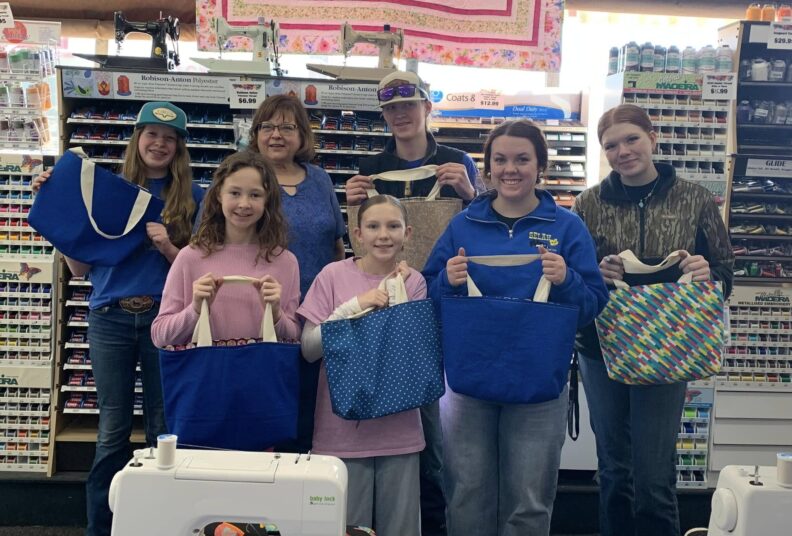 Seven people all smiling while standing in front of a display of sewing thread spools holding their handmade reversible fabric tote bags