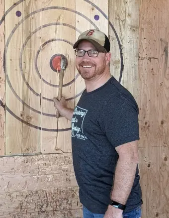 Light skinned middle aged man wearing black glasses, brown hat, smiling and standing for picture with right hand holding a pick axe that's stuck to a wooden target.