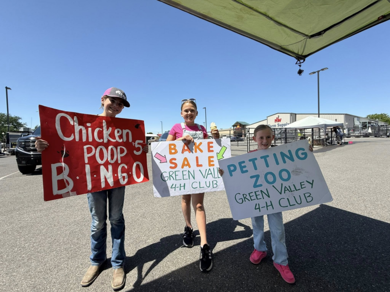 Three young girls holding signs advertising the different activities at their club fundraising event. Chicken poop bingo, bake sale, petting zoo for Green Valley 4-H Club