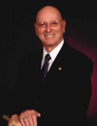 Light skinned older man wearing glasses and black suit with navy blue tie, smiling with hands crossed 