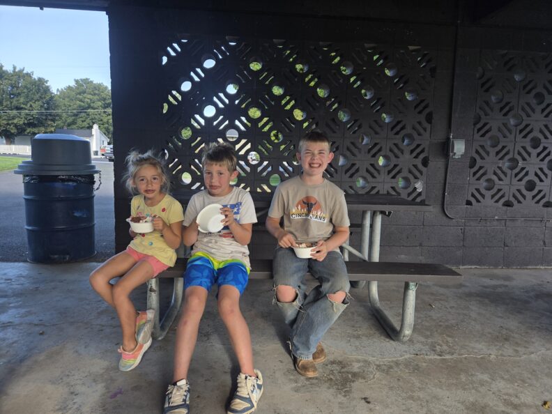 one female and two male youth members smiling eating ice cream while sitting at a park picnic table. 