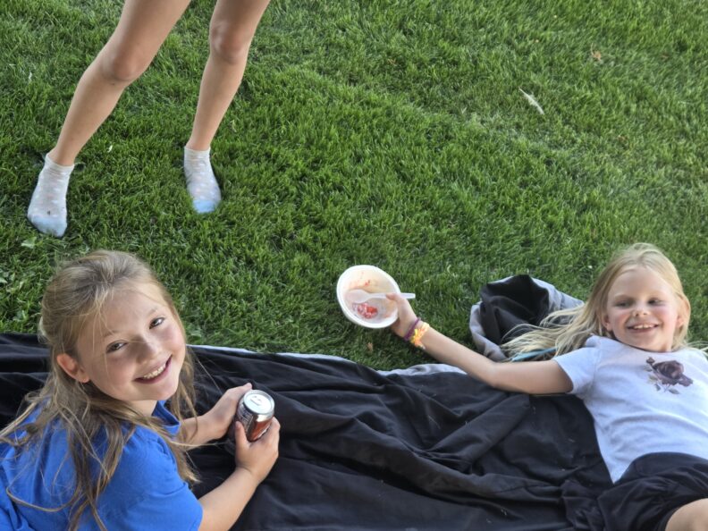 Two 8 year old female, both lying on a black blanket in the grass smiling. One holding a disposable ice cream bowl the other an aluminum can of soda.