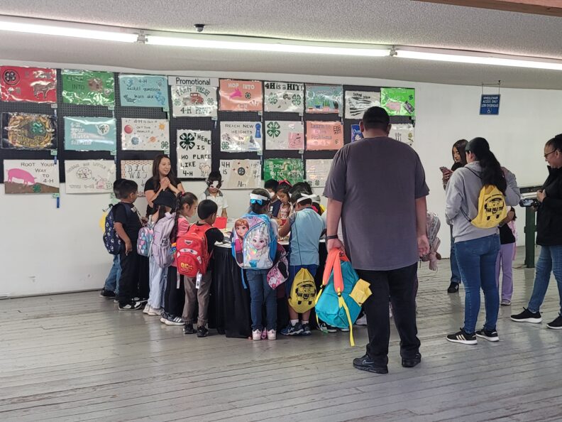 Group of young children wearing backpacks, gathered around a black table doing an activity with two adults at either end. At one end, a female wearing black smiles and clasps her hands together while speaking; at the other end, a male in a gray shirt, black pants, and shoes watches the children while holding a colorful backpack.