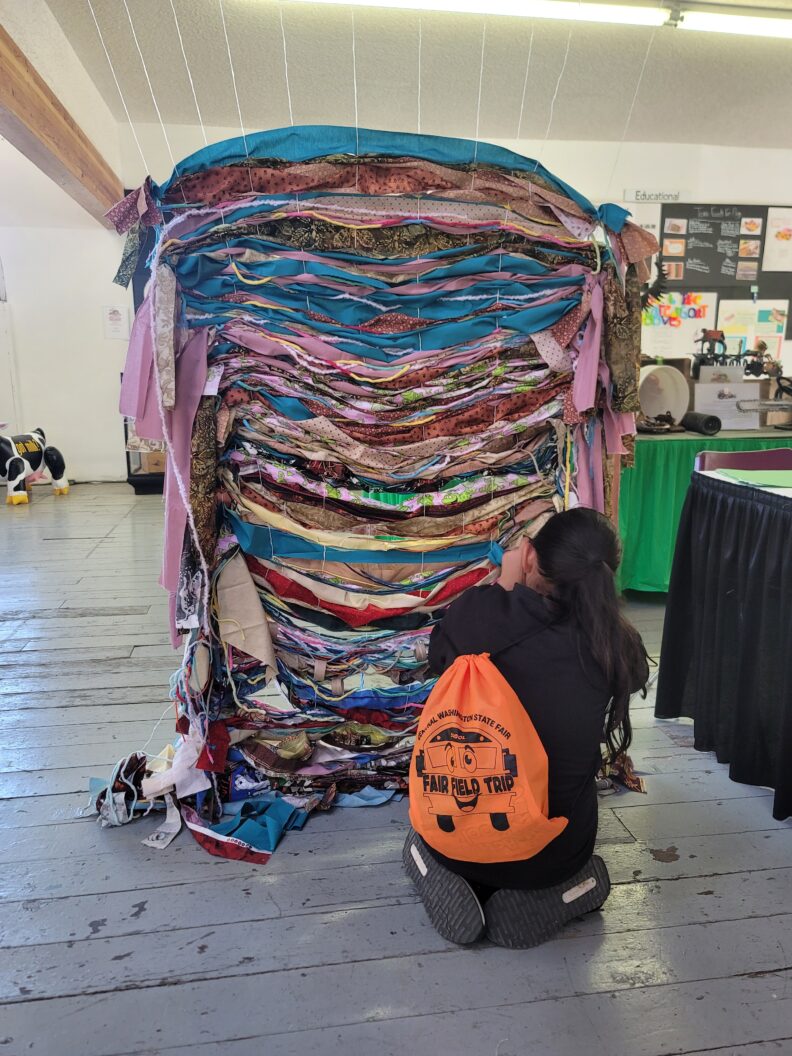 Youth with long dark hair, black attire, and an orange string backpack that states, "Fair Field Trip". Kneeling on the floor while assorting colorful material onto strings hanging from the ceiling to the floor to make a quilt.