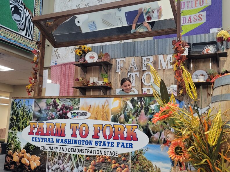 Girl smiling on the Farm to Fork Stage with a mirror above her reflecting the ingredients she will be using to make her dish.