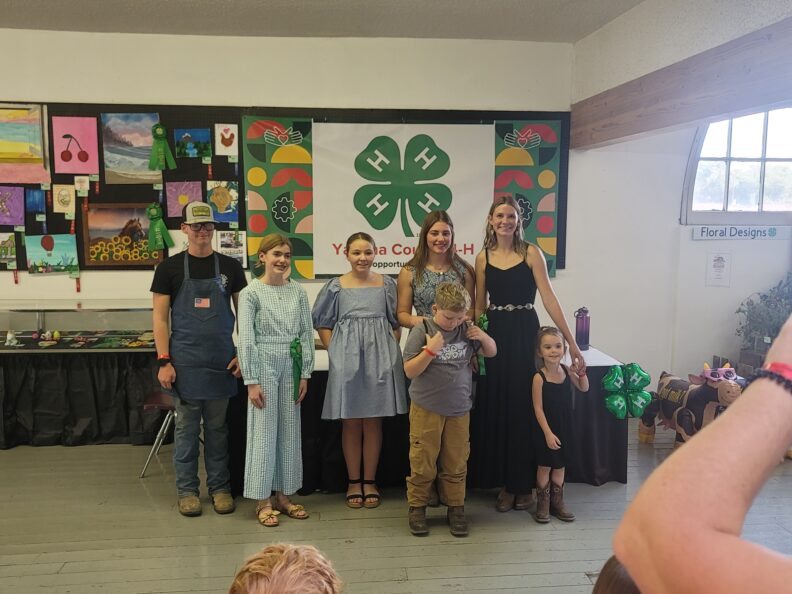Five girls and two boys, aged five to nineteen, are smiling in front of a "Yakima County 4-H" sign, showcasing clothes they made in the 4-H sewing project.