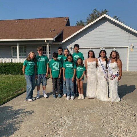 Eight youth in green "Harrah Humdingers 4-H club" shirts are smiling next to three girls in cream-colored dresses, sashes, and tiaras, all standing in the driveway of a gray house.