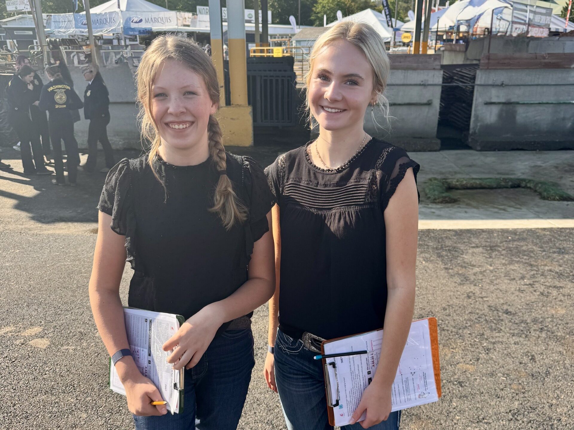 Two girls wearing black attire holding clipboards and paper smiling for a picture while waiting for the livestock judging contest to begin.