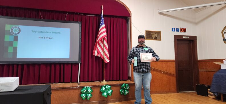 A man smiling in a ballcap and flannel shirt, while standing next to a small stage, proudly holding a certificate and thermal cup.
