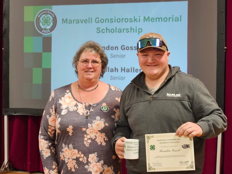 A smiling woman and a young person in a ballcap stand in front of a projector screen that states "Maravell Gonsioroski Memorial Scholarship". The young person is holding a certificate. and mug.